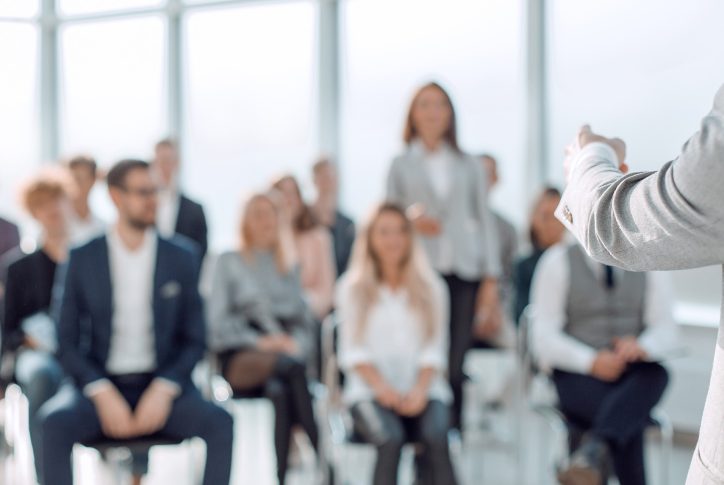 Das Publikum eines Vortrags ist verschwommen, scharfgestellt ist der Arm des Sprechers, der auf eine stehende Person des Publikums zeigt.